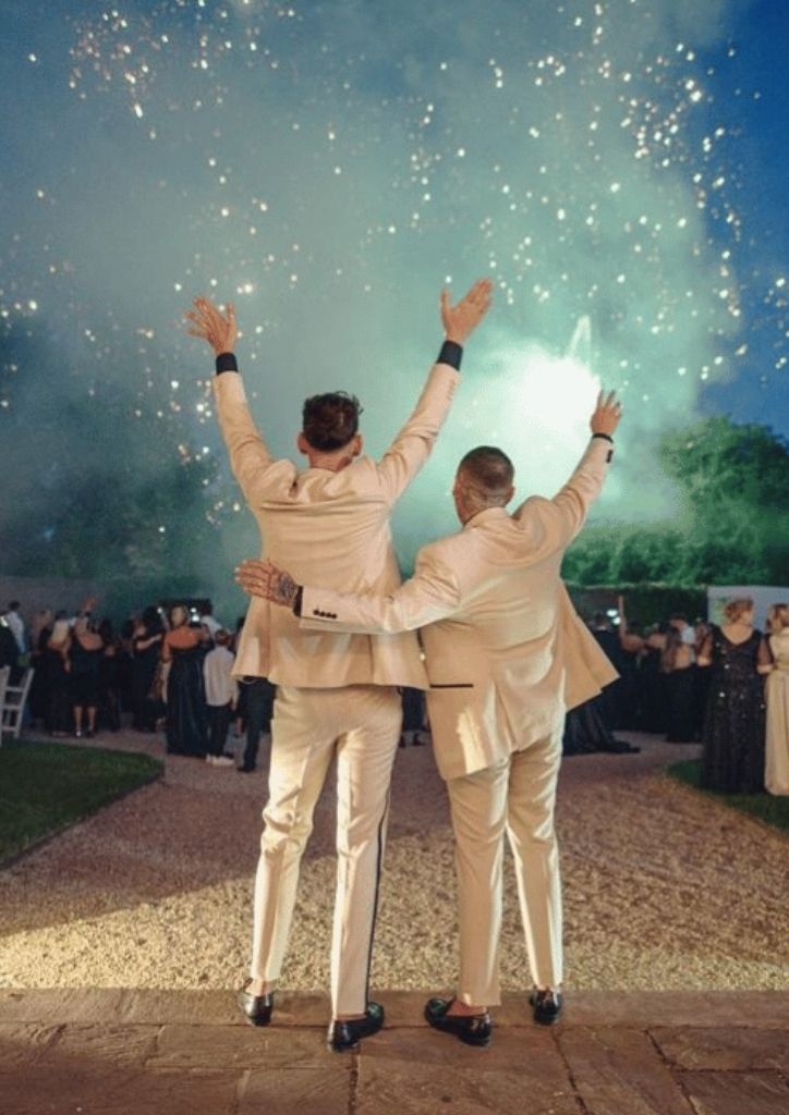 Newly-wed couple with their hands in the air, turned away from the camera, looking at the sparkles of fireworks and smoke in the background 
